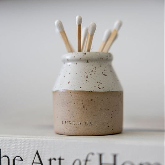 Small speckled ceramic jar with wooden sticks on top of a book titled 'The Art of Home'.