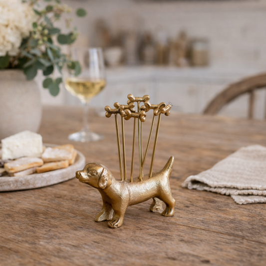 Gold dog-shaped toothpick holder on a wooden table with a glass of wine and snacks in the background.