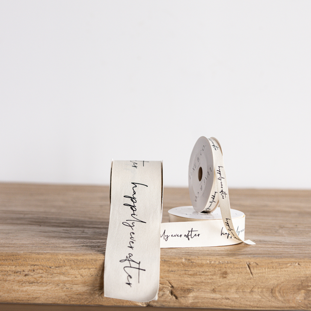 Cursive writing ribbon with "Happily Ever After" design by Luxe B Co Wholesale, featuring elegant script on white satin material.