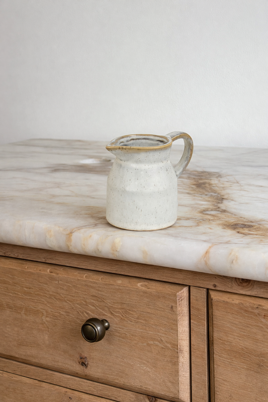 White ceramic pitcher with gold rim on a marble countertop next to wooden cabinets.